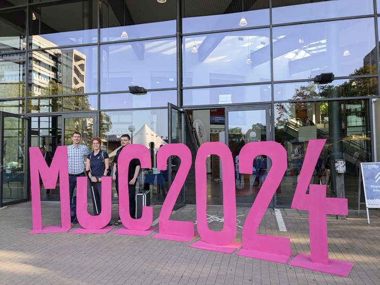 Der Schriftzug MuC2024 in pinken Holzbuchstaben vor einer Fensterfront.