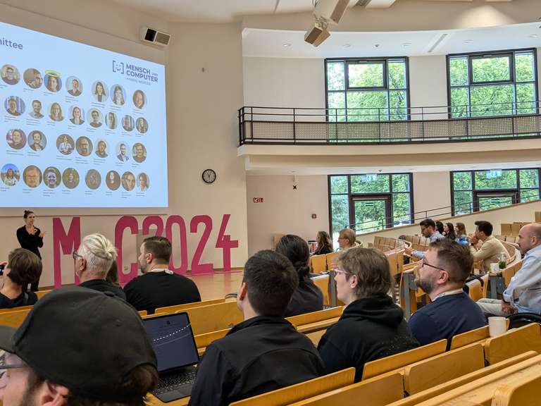 Viele Menschen sitzen in einem Saal in Reihen und schauen gebannt nach vorn.