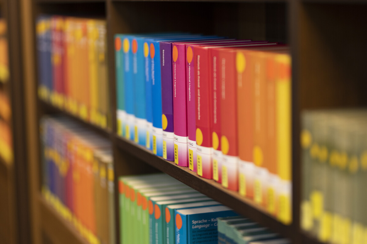 Ein Regal in einer Bibliothek. Vorn und Hinten sind die Bücher unscharf, dazwischen eine sortierte Sammlung farblich von Orange über Rot und Violett zu Blau abgestuft.