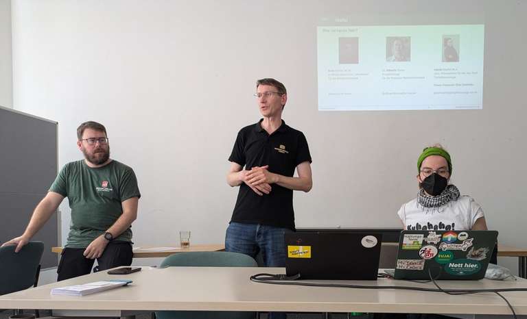 Andy, Albrecht und Karola vorn in einem Raum. Davor ein Workshoptisch, dahinter klein eine Präsentationsfolie