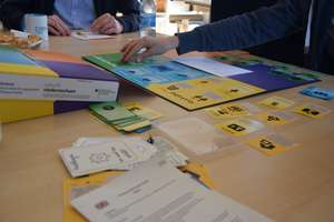 Auf einem Tisch ausgebreitet ein Spielbrett mit großen Feldern, in denen verschiedene Karten liegen; von einer Person wird eine neue Karte dazugelegt