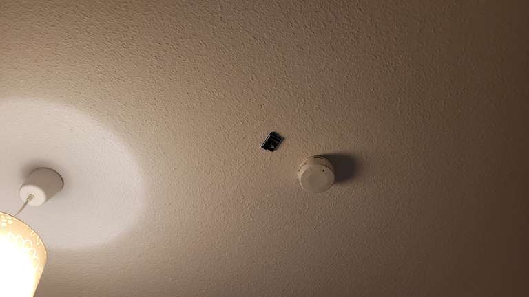 neben einem Rauchmelder klebt ein Sensortag des Sensorkits an der Zimmerdecke; in der einen Ecke des Bildes ist eine Lampe zu sehen