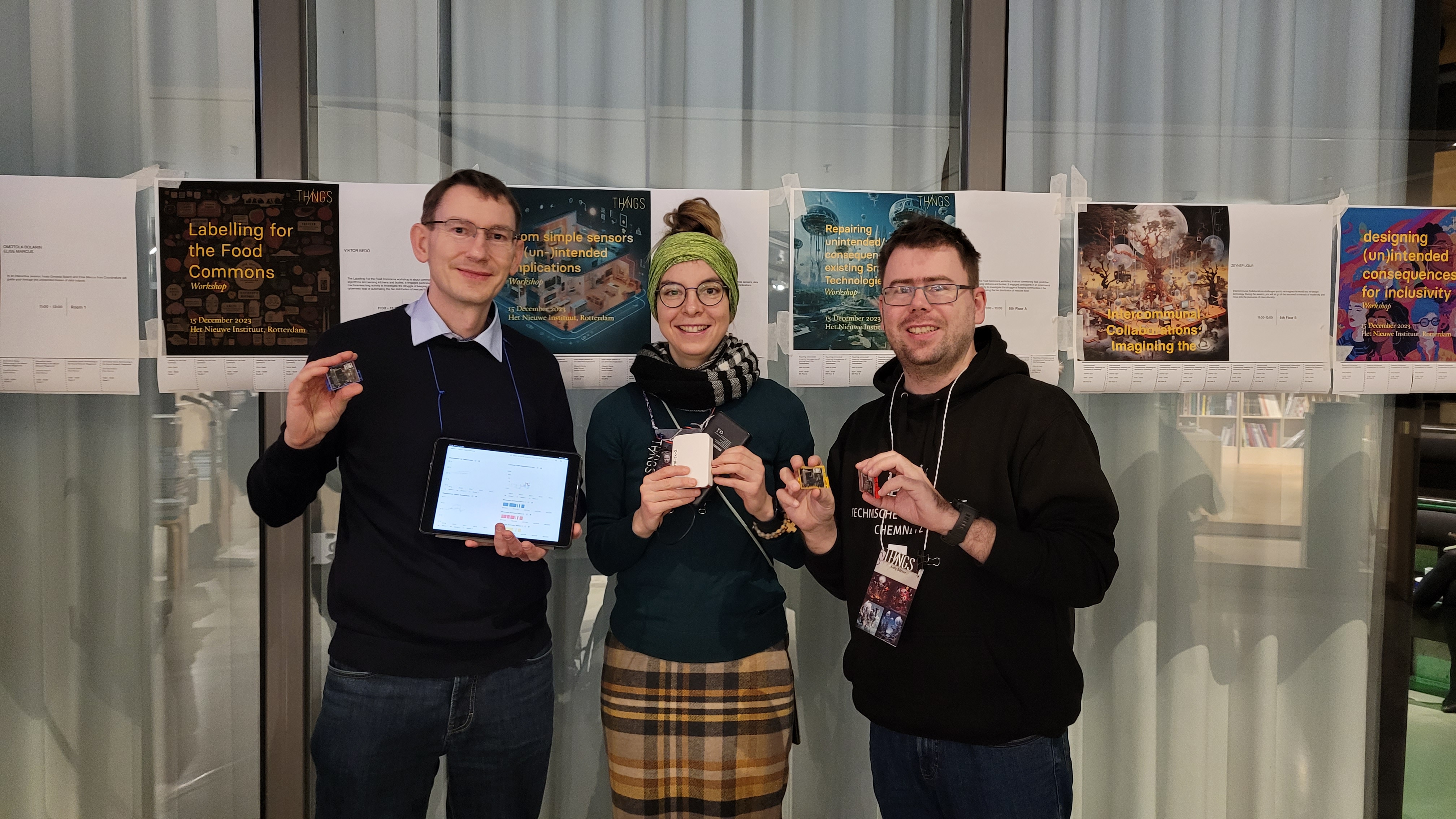 Albrecht, Karola und Andy auf der ThingsCon23. Sie halten ein Tablet, einen Mini-Computer und drei Sensortags in den Händen. Im Hintergrund Poster der Workshops.