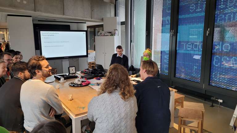 Vorstellungsrunde unter den Workshop-Teilnehmern. Diese sitzen um einen Tisch herum, im Hintergrund die Präsentation für den Workshop