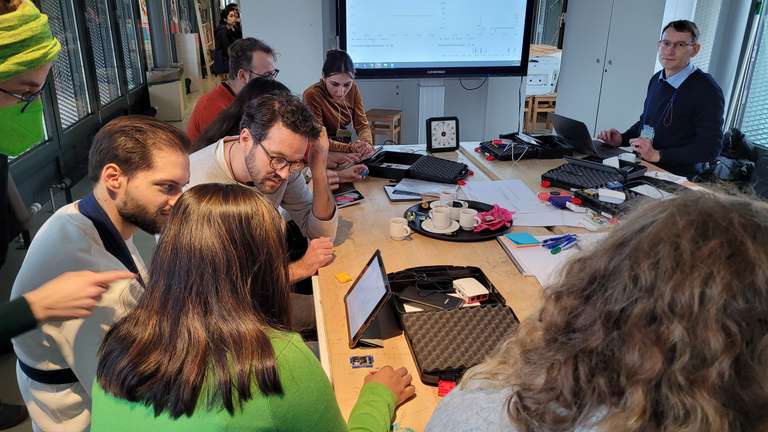 Blick in den Workshopraum. Es sind mehrere Personen zu sehen und im Hintergrund ein Fernseher.