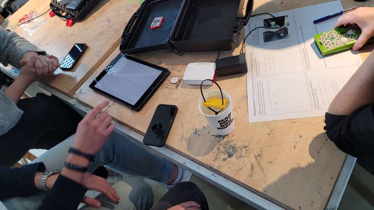 Auf einem Tisch sind Inhalt des Sensorkits und Canva für den Workshop ausgebreitet. In einem leeren Kaffeebecher hängt das gelbe  Sensortag. Um den Tisch herum mehrere Arme von Workshopteilnehmern bei der Zusammenarbeit und Diskussion